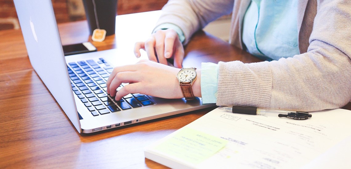 person sat at desk with coffee researching on laptop with a notebook