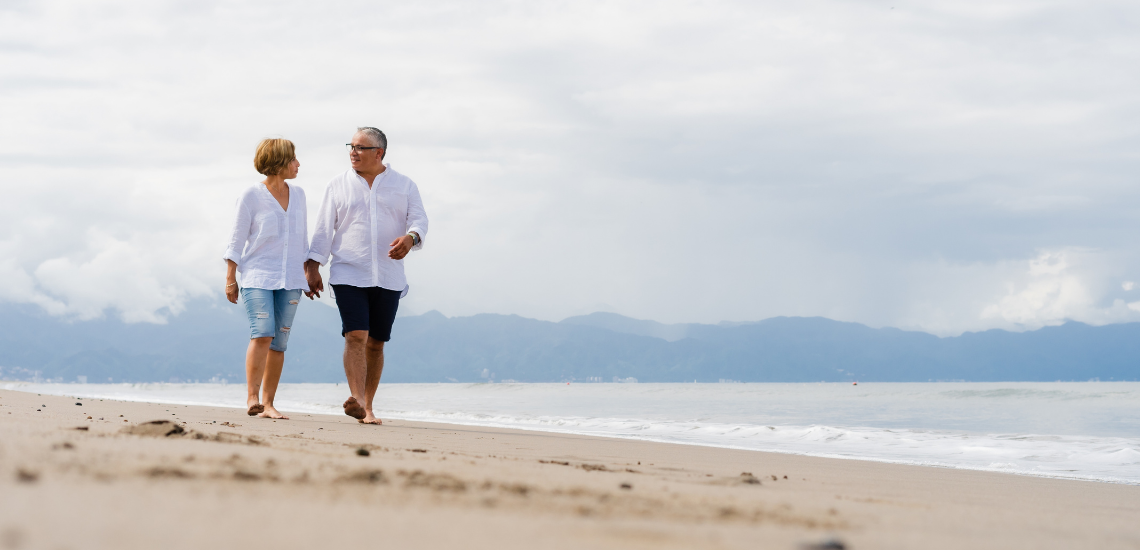 Older couple walking barefoot along a sandy beach, smiling and talking together.